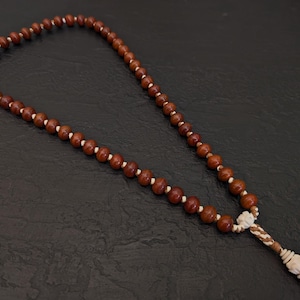 May include: A strand of prayer beads with dark brown wooden beads and white spacers. The beads are strung together with a white and brown braided cord, ending in a decorative tassel with a carved ivory-colored element. The beads are arranged on a dark surface.