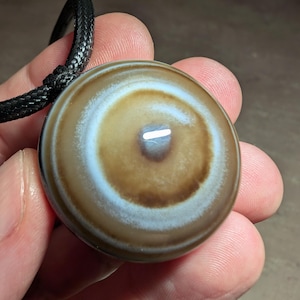 May include: A circular, polished stone pendant featuring concentric bands of brown, white, and dark brown, resembling an eye pattern. The pendant is suspended from a black braided cord, showcasing a smooth, glossy surface with a reflective highlight.