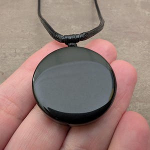 K&ouml;nnte beinhalten: Ein schwarzer Obsidian-Anh&auml;nger an einer schwarzen Schnurkette. Der Anh&auml;nger ist rund und glatt.