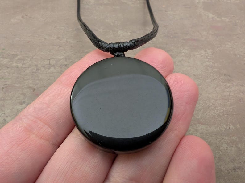 K&ouml;nnte beinhalten: Ein schwarzer Obsidian-Anh&auml;nger an einer schwarzen Schnurkette. Der Anh&auml;nger ist rund und glatt.