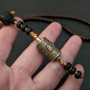 May include: A black and orange beaded necklace with a silver prayer wheel pendant. The prayer wheel has intricate carvings and the word "Om" is visible.