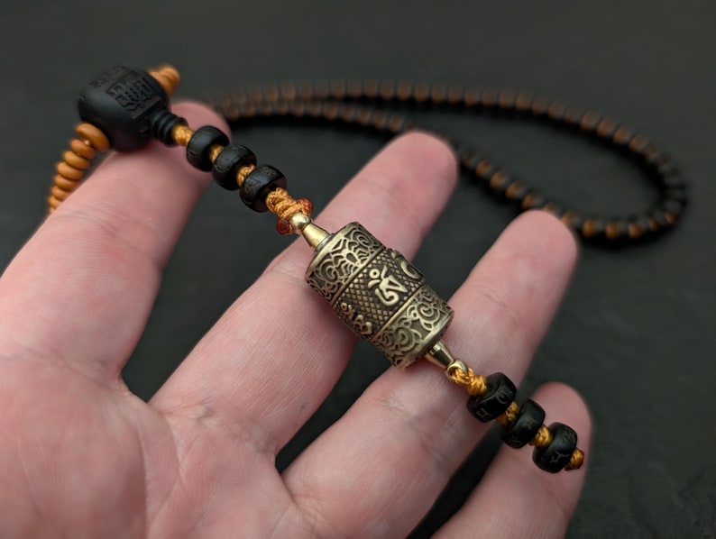 May include: A black and orange beaded necklace with a silver prayer wheel pendant. The prayer wheel has intricate carvings and the word "Om" is visible.
