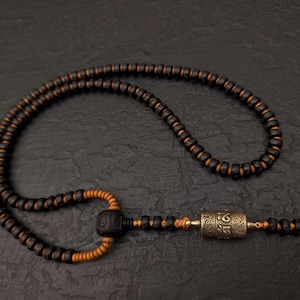 May include: A black and brown wooden mala necklace with a silver prayer wheel pendant. The necklace is made of small beads and has a tassel at the end.
