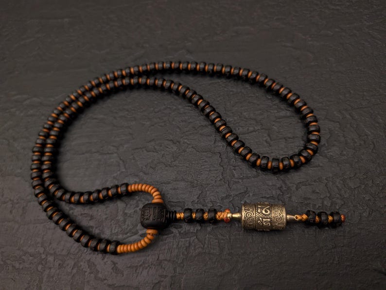 May include: A black and brown wooden mala necklace with a silver prayer wheel pendant. The necklace is made of small beads and has a tassel at the end.