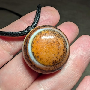Puede incluir: Collar con colgante redondo y cordón negro. El colgante presenta una mezcla de colores naranja, marrón y blanco. El colgante tiene un diseño circular y una superficie texturizada.