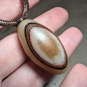 May include: An oval-shaped pendant with a smooth, polished surface featuring concentric rings of cream, tan, and dark brown colors. The pendant is suspended from a dark brown, textured, braided cord that loops through the top of the stone.