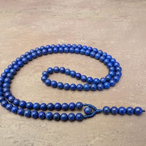May include: A long strand of deep blue lapis lazuli beads arranged in a circular pattern. The necklace features a series of uniform, polished spherical stones strung together, ending in a knotted section with a single larger bead and a small tassel.