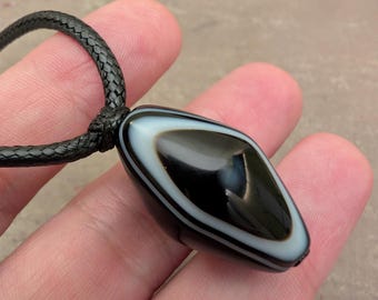 May include: A black and white banded agate pendant with a smooth, polished, diamond-like shape. The pendant is suspended from a black braided cord necklace, featuring a distinct eye-like pattern with a dark center surrounded by a white ring.