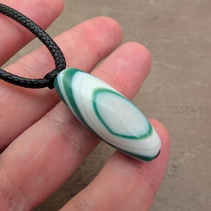 May include: A cylindrical pendant featuring a smooth, polished surface with concentric bands of emerald green and opaque white. The pendant is suspended from a black braided cord, creating a simple necklace design with a glossy, stone-like finish.