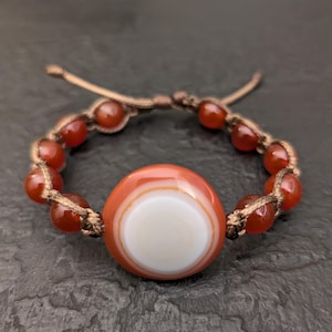 May include: A bracelet featuring a large, round, orange and white banded agate focal bead centered among smaller, smooth, translucent orange carnelian beads. The beads are strung on a brown cord with an adjustable sliding knot closure mechanism.