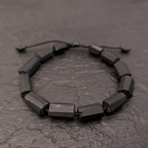 Puede incluir: Una pulsera de cuentas gris oscuro con cierre de nudo corredizo. La pulsera presenta múltiples cuentas de forma geométrica, espaciadas uniformemente. Las cuentas tienen un acabado mate y están ensartadas en un cordón oscuro. La pulsera se muestra sobre una superficie oscura y texturizada.