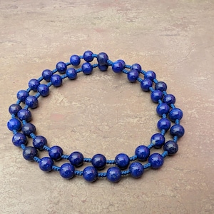 May include: A double-strand necklace featuring round, deep blue lapis lazuli beads with visible natural mineral inclusions. The beads are strung on a matching blue cord with small, hand-tied knots between each individual stone bead.