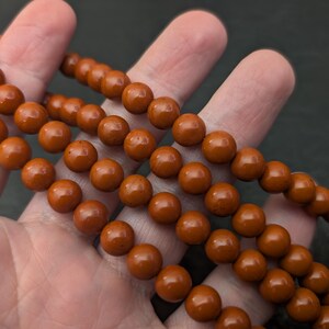 May include: A strand of smooth, round, rust-colored beads. The beads are uniform in size and shape, featuring a polished, glossy finish that reflects light. The strand is arranged in multiple loops, showcasing the consistent deep orange-brown color.
