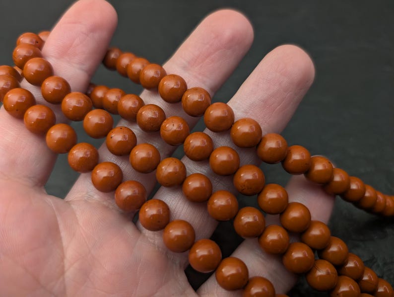 May include: A strand of smooth, round, rust-colored beads. The beads are uniform in size and shape, featuring a polished, glossy finish that reflects light. The strand is arranged in multiple loops, showcasing the consistent deep orange-brown color.