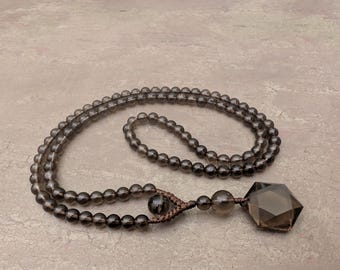 May include: A necklace featuring a long strand of translucent, smoky gray spherical beads. The strand is connected by brown cord to a larger, faceted, hexagonal smoky gray gemstone pendant, accented by two additional round beads near the pendant.