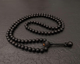 May include: A black wooden beaded necklace with a decorative pendant and tassel. The necklace is made of small, round beads and is displayed on a dark textured surface. The pendant has intricate carvings.