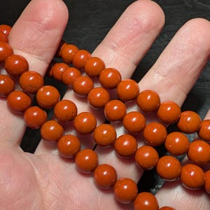 May include: A strand of smooth, round, opaque burnt-orange beads. The beads are uniform in size and shape, featuring a consistent matte to semi-gloss finish with subtle natural variations in the deep orange color throughout the entire string.