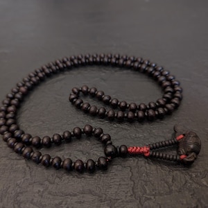 Può includere: Una collana di perline marrone scuro con un ciondolo decorativo. La collana è composta da piccole perline rotonde infilate insieme. Il ciondolo presenta un design intricato ed è collegato alla collana con filo rosso.