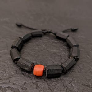 Puede incluir: Una pulsera de cuentas negras con una sola cuenta naranja. La pulsera está hecha de cuentas oscuras de forma geométrica ensartadas en un cordón negro. La cuenta naranja añade un toque de color al diseño monocromático.