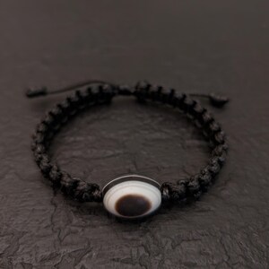 May include: Black macrame bracelet featuring a central bead with a white and brown eye-like design. The bracelet is adjustable with sliding knots. The bead has a dark brown center surrounded by a white band.