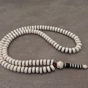 May include: A white prayer bead necklace with a black and white tassel. The beads are round and have a smooth finish.