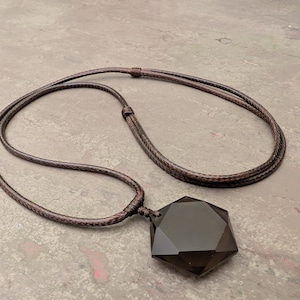 Peut inclure: Un collier avec un cordon en cuir brun foncé et un grand pendentif en pierre noire facettée. Le pendentif a une forme géométrique et réfléchit la lumière. Le cordon tressé est réglable, pour un style simple et élégant.