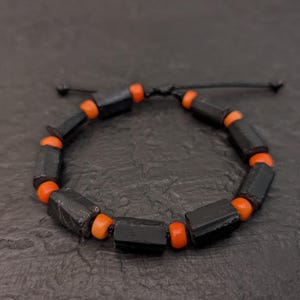 Puede incluir: Pulsera ajustable con cuentas geométricas negras y cuentas naranjas. La pulsera está ensartada en un cordón negro, creando un contraste llamativo. El diseño es simple pero llamativo, adecuado para el uso diario.