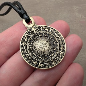 May include: A bronze pendant necklace with a detailed circular design. The pendant features intricate patterns and symbols, suspended from a black cord. The design includes animal figures and a central geometric pattern.