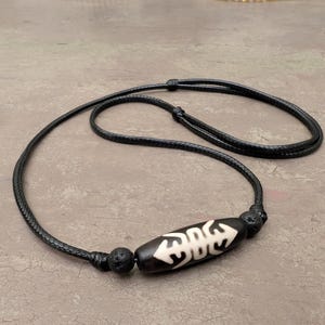 May include: Black cord necklace featuring a central, oblong bead with a black and white tribal design. The necklace has adjustable length and includes two round, textured beads on either side of the central bead.