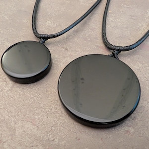 K&ouml;nnte beinhalten: Zwei schwarze Obsidian-Anh&auml;ngerketten mit schwarzen Schn&uuml;ren. Der gr&ouml;&szlig;ere Anh&auml;nger hat einen Durchmesser von etwa 5 cm, der kleinere einen Durchmesser von etwa 2,5 cm.