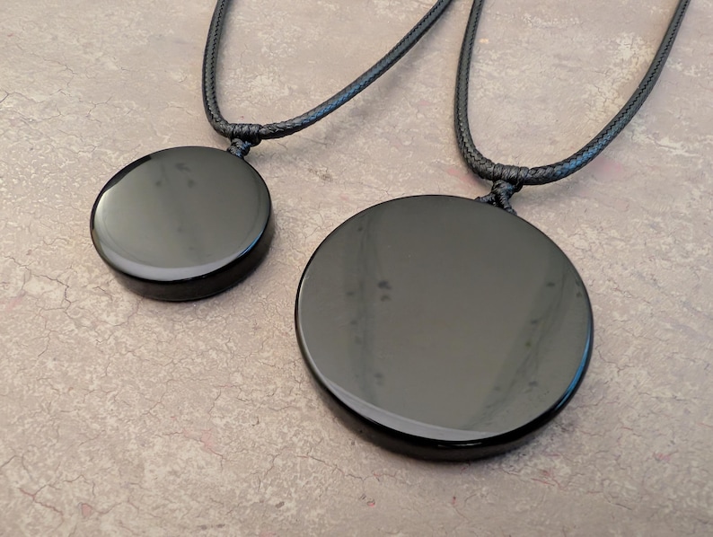 K&ouml;nnte beinhalten: Zwei schwarze Obsidian-Anh&auml;ngerketten mit schwarzen Schn&uuml;ren. Der gr&ouml;&szlig;ere Anh&auml;nger hat einen Durchmesser von etwa 5 cm, der kleinere einen Durchmesser von etwa 2,5 cm.