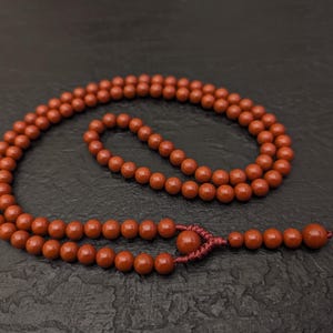 May include: A strand of round, matte, terracotta-colored beads arranged in a circular loop. The beads are strung on a matching dark red cord, featuring a decorative knot detail that secures a small cluster of additional beads at the end of the strand.