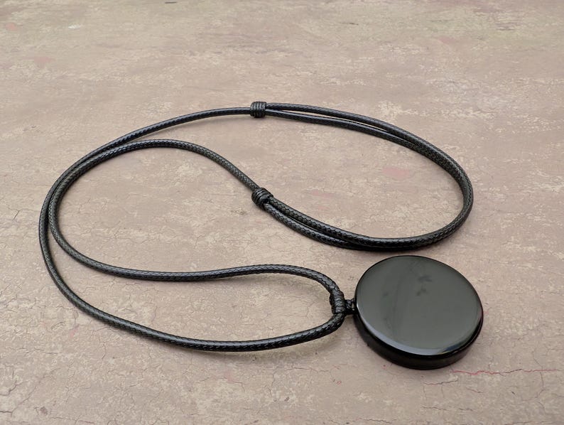 K&ouml;nnte beinhalten: Eine schwarze Halskette mit einem runden, glatten schwarzen Steinanh&auml;nger. Die Halskette besteht aus einer schwarzen Schnur.
