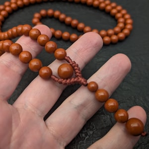 May include: A strand of smooth, round, terracotta-colored beads strung on a matching brown cord. The necklace features a central knot detail with a larger bead positioned above a smaller dangling section of beads, creating a continuous loop.
