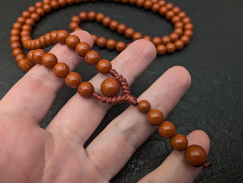 May include: A strand of smooth, round, terracotta-colored beads strung on a matching brown cord. The necklace features a central knot detail with a larger bead positioned above a smaller dangling section of beads, creating a continuous loop.