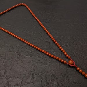 May include: A necklace featuring a long strand of small, round, terracotta-colored beads. The beads are strung on a matching red cord, which includes a decorative knotted section near the bottom that leads to a short, dangling extension of beads.