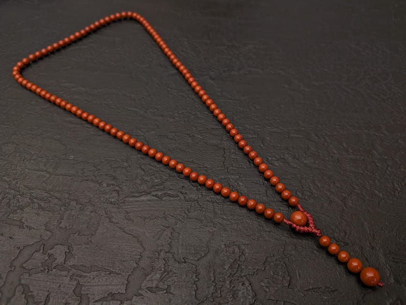 May include: A necklace featuring a long strand of small, round, terracotta-colored beads. The beads are strung on a matching red cord, which includes a decorative knotted section near the bottom that leads to a short, dangling extension of beads.