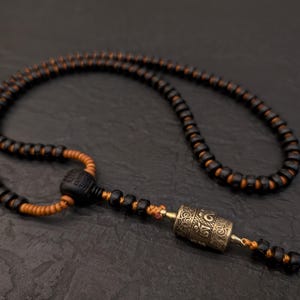 May include: A black and orange beaded necklace with a silver prayer wheel pendant. The prayer wheel has intricate designs and is attached to the necklace with a black cord.