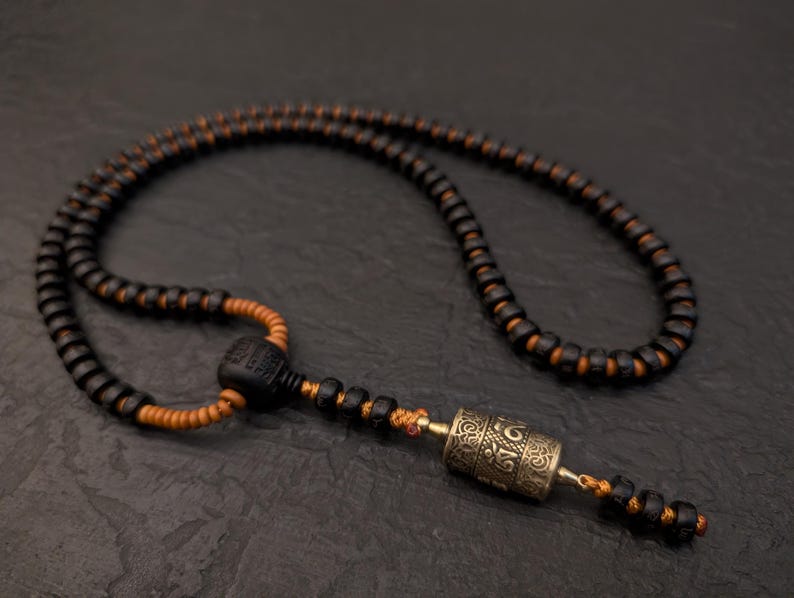 May include: A black and orange beaded necklace with a silver prayer wheel pendant. The prayer wheel has intricate designs and is attached to the necklace with a black cord.