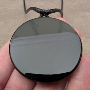 K&ouml;nnte beinhalten: Ein schwarzer Obsidian-Anh&auml;nger an einer schwarzen Schnurkette. Der Anh&auml;nger ist rund und glatt, mit einem hohen Glanz.