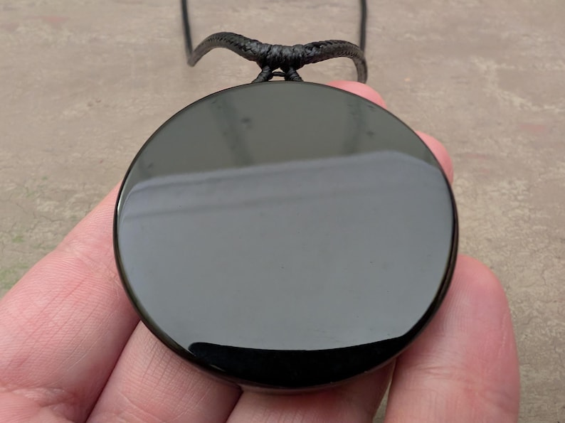 K&ouml;nnte beinhalten: Ein schwarzer Obsidian-Anh&auml;nger an einer schwarzen Schnurkette. Der Anh&auml;nger ist rund und glatt, mit einem hohen Glanz.