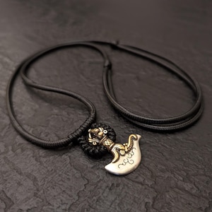 May include: A silver and gold pendant necklace with a black cord. The pendant features an ornate design with a curved blade shape. The necklace is displayed on a dark, textured surface.