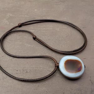 Puede incluir: Un collar con un colgante de piedra redonda, lisa y de color blanco translúcido con un centro marrón oscuro, suspendido de un cordón trenzado de color marrón oscuro. El cordón incluye nudos deslizantes ajustables para modificar la longitud.