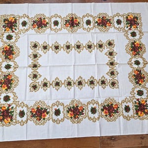 May include: A rectangular white tablecloth with a floral design. The border and corners feature ornate floral arrangements in shades of red, yellow, and brown, framed by a gold-colored decorative trim.