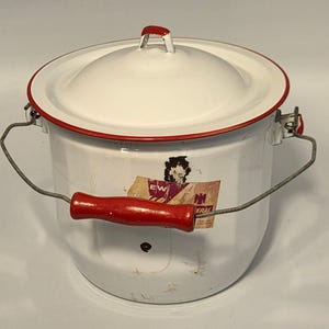 May include: White enamelware pot with a lid and a red handle. The pot has a red rim and a metal handle. A faded sticker is on the side of the pot. The pot is likely vintage and used for cooking or storage.