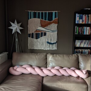 May include: A pink braided decorative pillow rests on a tan leather couch. A woven wall hanging with blue, brown, and white accents hangs above the couch. A white star-shaped lamp and a bookshelf are also visible.
