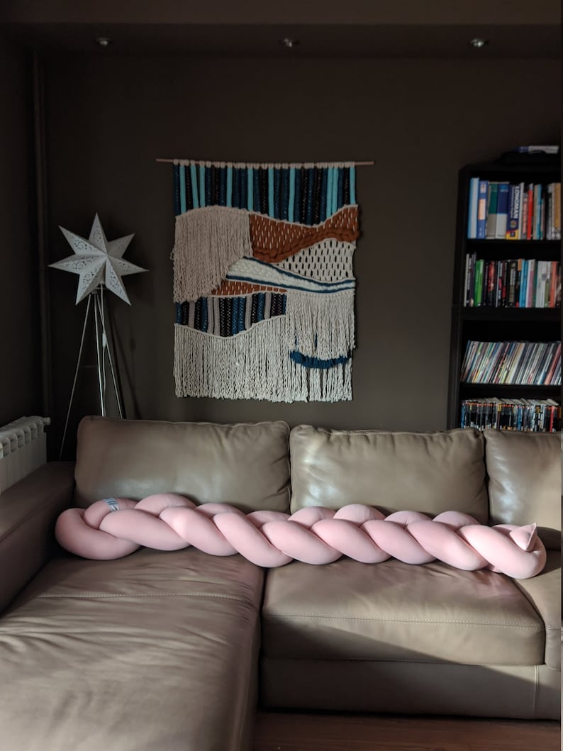 May include: A pink braided decorative pillow rests on a tan leather couch. A woven wall hanging with blue, brown, and white accents hangs above the couch. A white star-shaped lamp and a bookshelf are also visible.