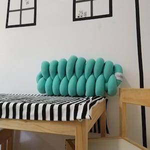 Op de afbeelding: Een gevlochten turquoise kussen ligt op een houten tafel bedekt met een zwart-wit gestreepte doek. Het kussen staat tegen een witte muur met zwarte venstercontouren. Rechts staat een houten stoel.