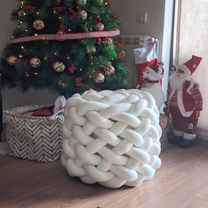 Könnte beinhalten: Ein weißer, geflochtener Pouf mit Korbgeflecht-Design. Der runde, würfelförmige Hocker ist aus dickem, weichem Material gefertigt. Ein Weihnachtsbaum, eine Weihnachtsmannfigur und andere Weihnachtsdekorationen befinden sich im Hintergrund.