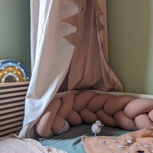 May include: A children's bedroom scene featuring a rainbow-shaped night light with warm white lights. A braided bumper surrounds the bed, and a small sheep figurine sits near a blanket with sheep patterns. A canopy hangs above the bed.